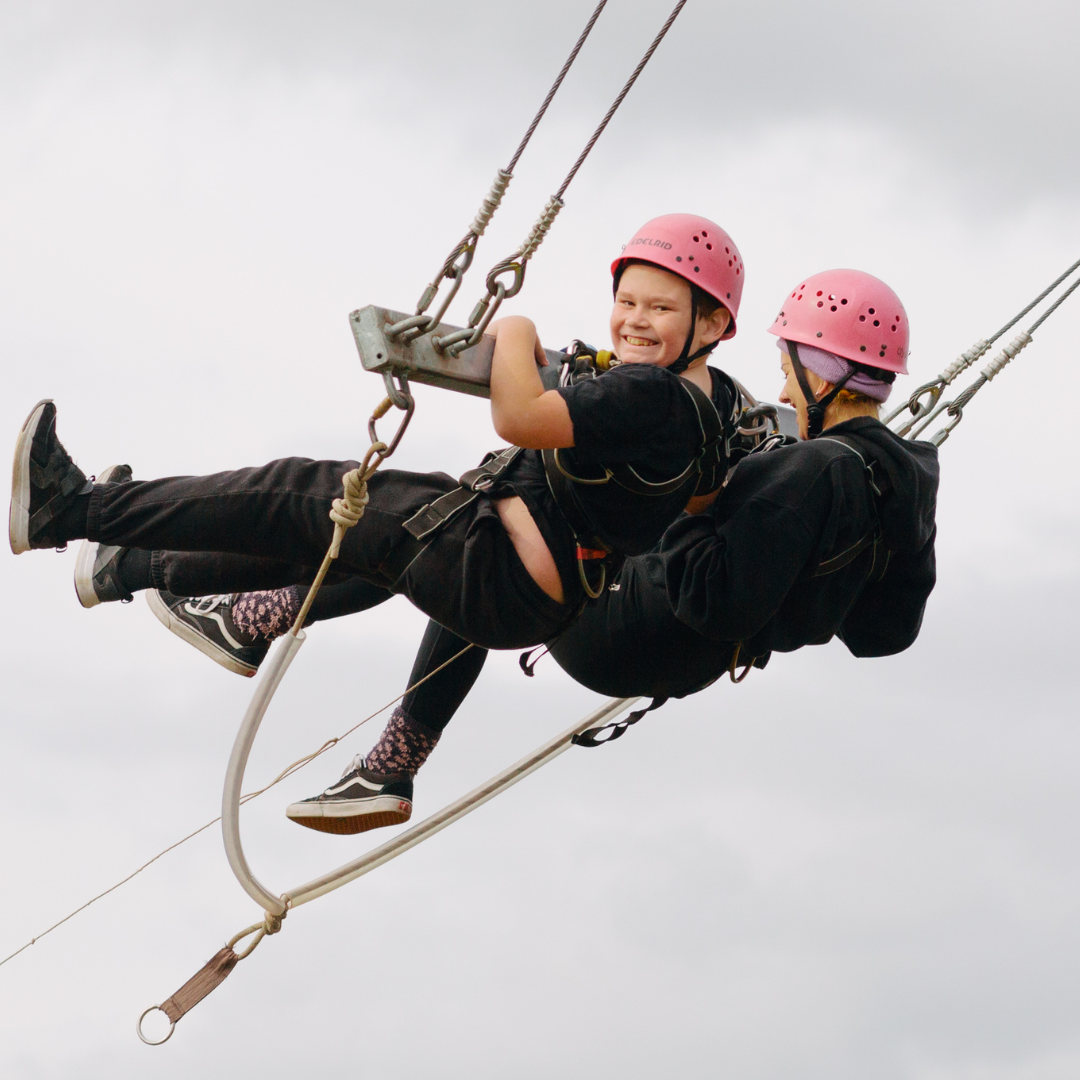 Two people in safety gear on a giant swing, one grinning at the camera